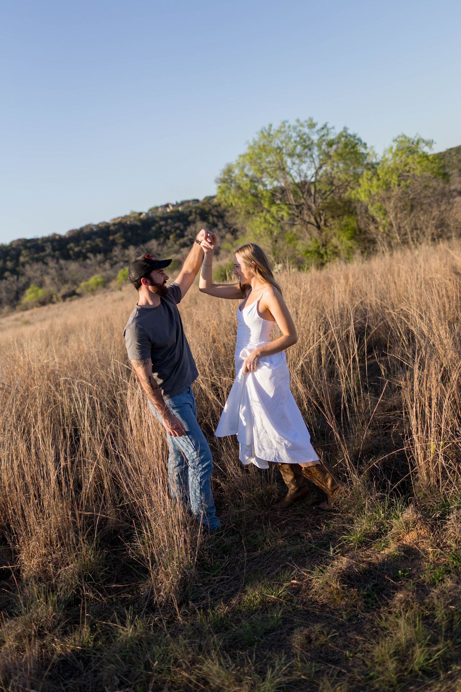 Couple dancing in golden field, man in cap and jeans spinning woman in white dress and cowboy boots at sunset.