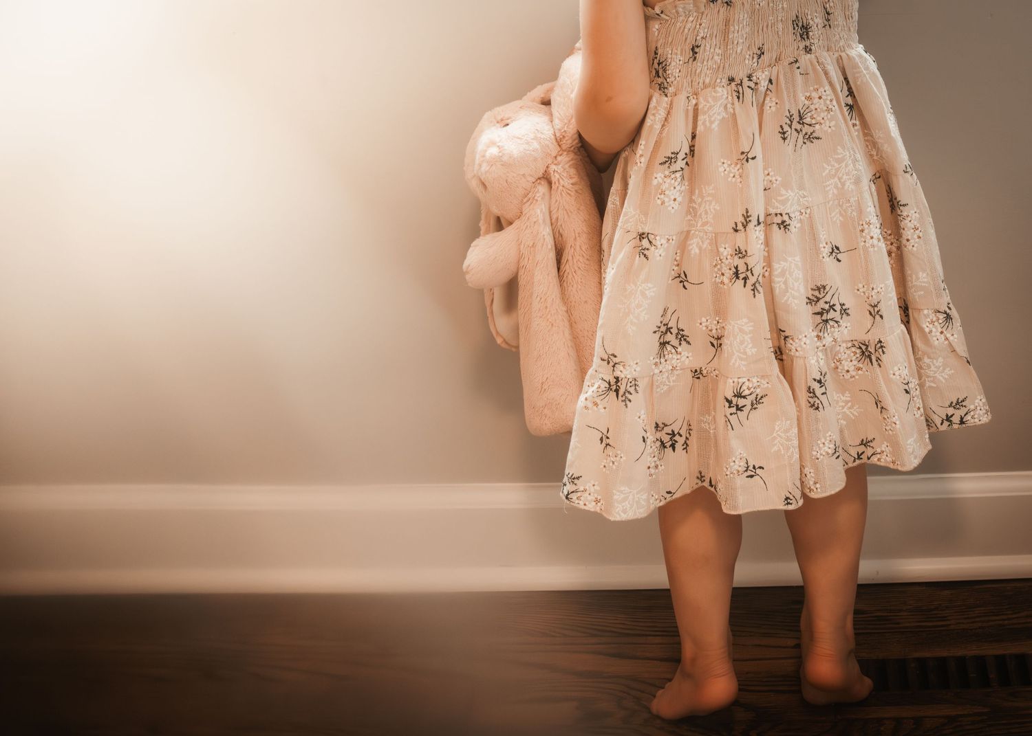 Young girl in floral dress holding a teddy bear, standing barefoot against a white wall on dark hardwood floor.