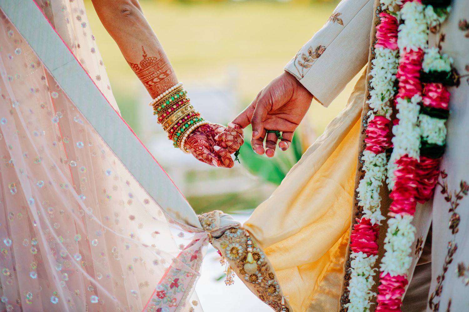 Indian bride and groom holding hands adorned with mehndi, bangles, and floral garlands in traditional wedding attire.