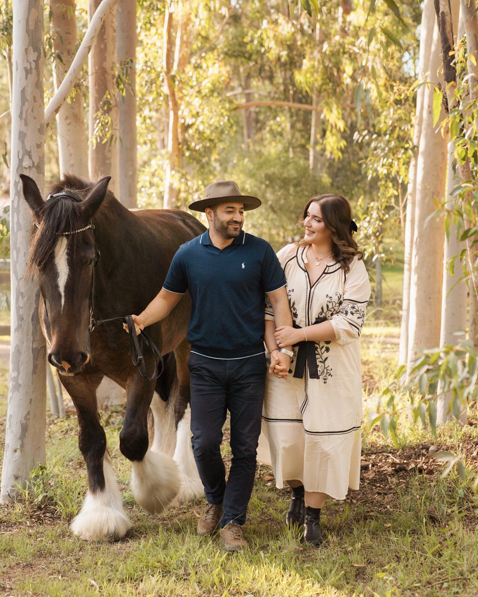 Couple walking with a horse through eucalyptus forest, man in hat and woman in cream dress during couples photoshoot with a horse in Sydney