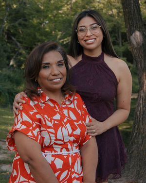 Two women smile outdoors among trees, one in a red floral dress, the other in a maroon halter dress with glasses.