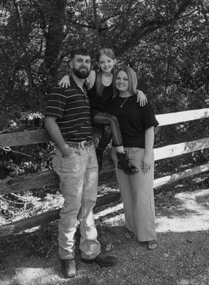 Black and white family portrait of two adults and a child posing by a wooden fence surrounded by trees.