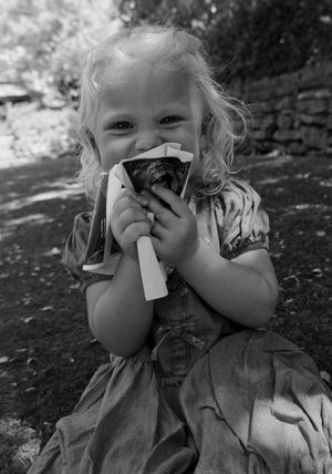 Smiling blonde toddler girl in a dress holding a small animal close to her face outdoors in black and white.