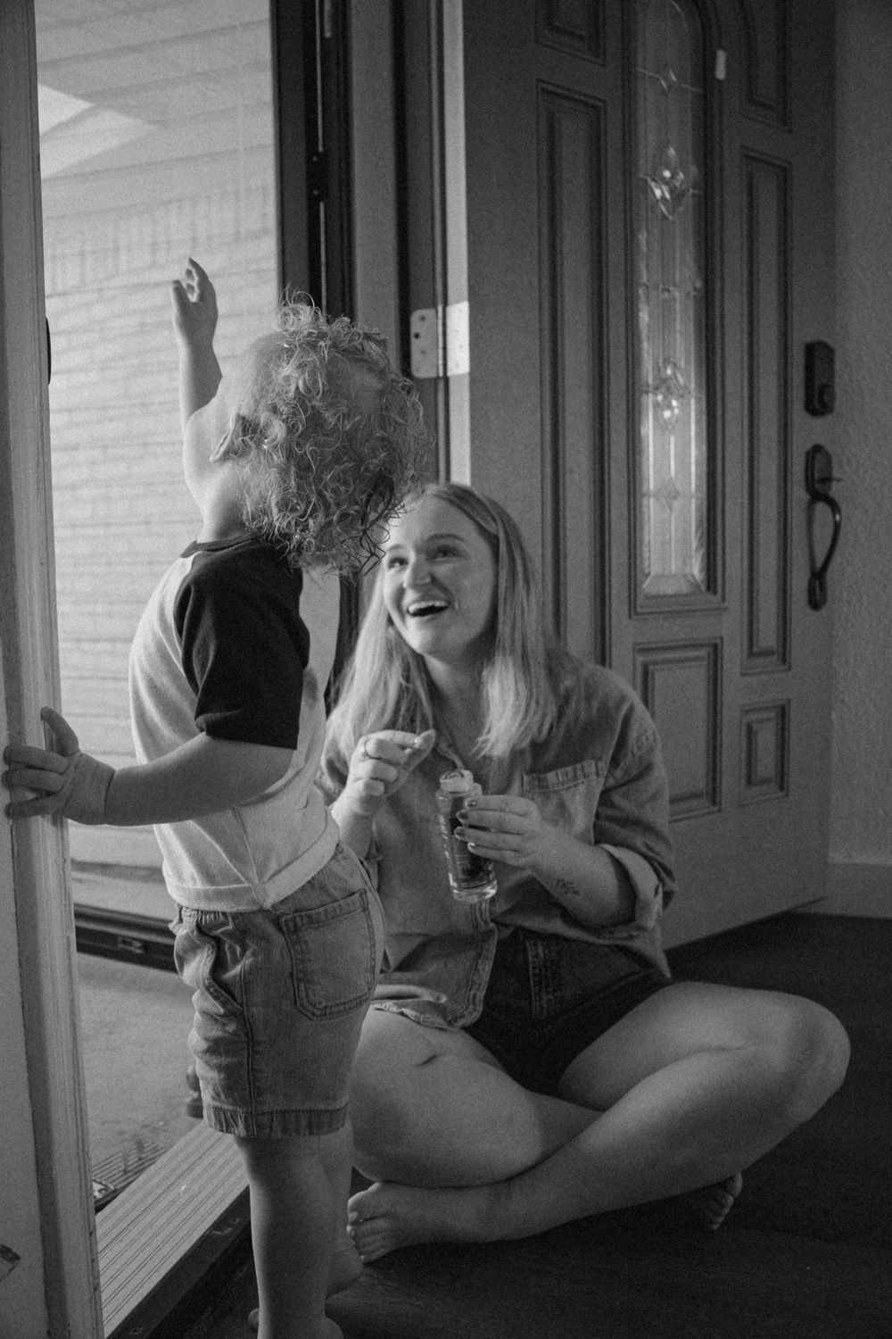 Black and white photo of a laughing woman crouching beside a curly-haired toddler at a front door.
