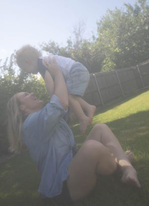 A woman lies on green grass in a backyard, joyfully lifting a toddler above her head on a sunny day.