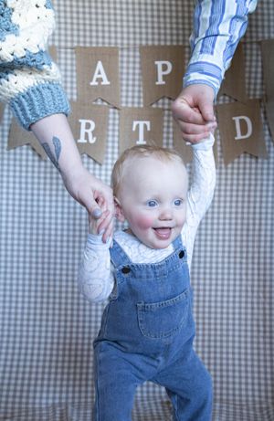 A smiling baby in denim overalls holds adults hands standing before a Happy Birthday banner on gingham background.