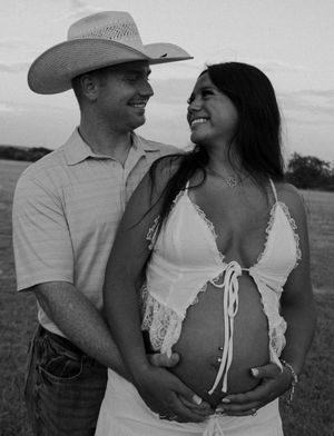 Black and white photo of a smiling pregnant couple outdoors, man wearing cowboy hat embracing woman in white lace dress.