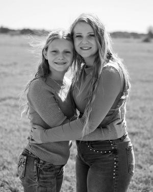Two smiling girls with long hair embrace outdoors in a black and white portrait photo in an open field.