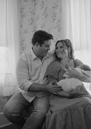Black and white photo of a couple smiling at each other while holding their newborn baby near a window.