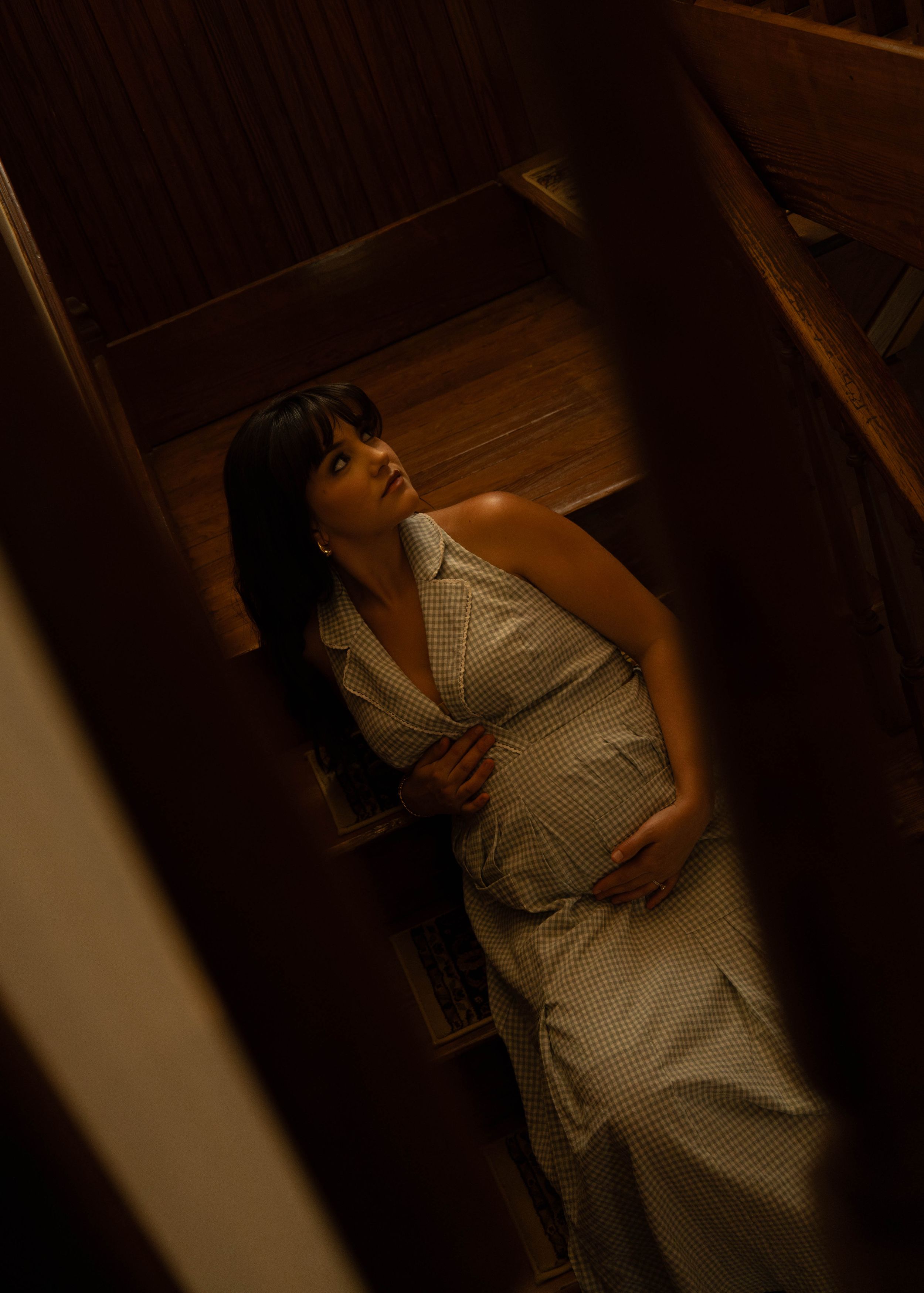 Pregnant woman in checkered dress standing in wooden closet, looking up with hands on belly in dim warm light.