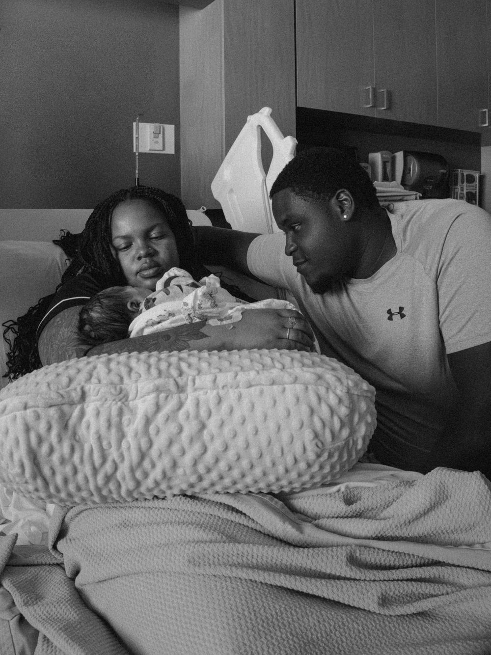 Black and white photo of new parents in hospital room admiring their newborn baby wrapped in blanket.
