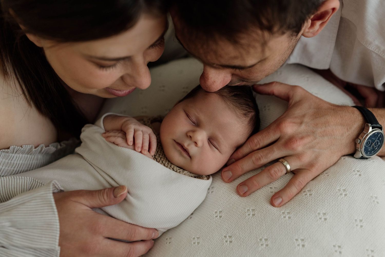 A couple smiles at their newborn baby on a bed with green walls and a New Zealand poster in a cozy bedroom.