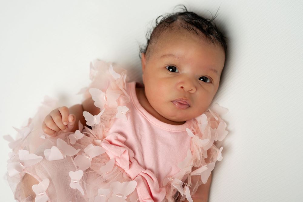 Newborn baby girl in pink ruffled outfit lying on white background, looking at camera with dark hair and bright eyes.