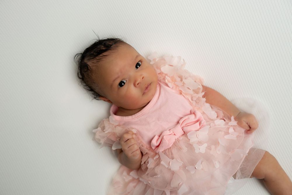 Cute newborn baby girl posed in a soft pink ruffle dress on a white surface.