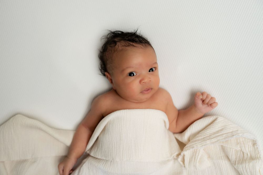 Newborn baby wrapped in cream muslin cloth lying on white background looking at camera.