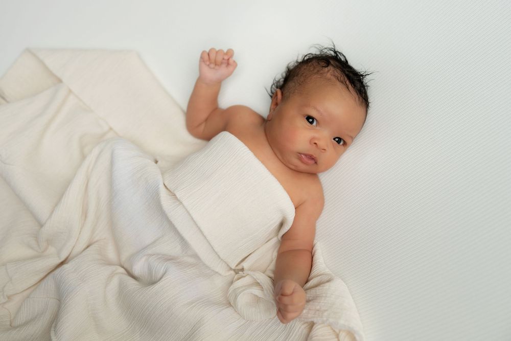 Newborn baby wrapped in cream muslin blanket lying on white background.