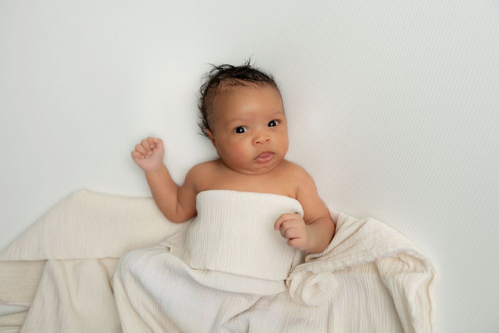 Newborn baby wrapped in a white swaddle blanket lying on a white background.