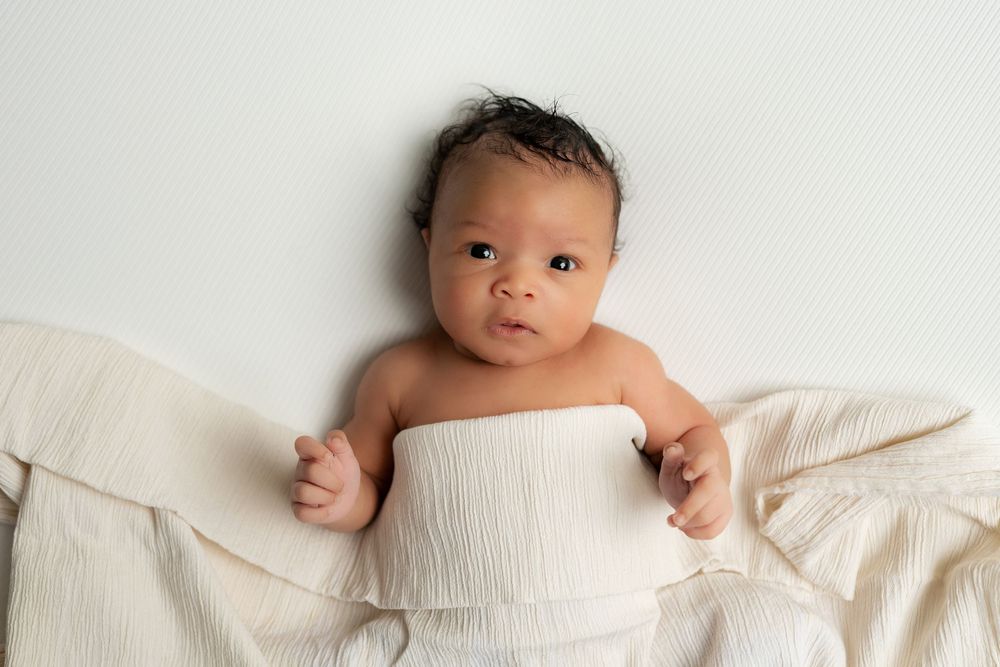 Newborn baby swaddled in white muslin wrap lying on cream blanket.