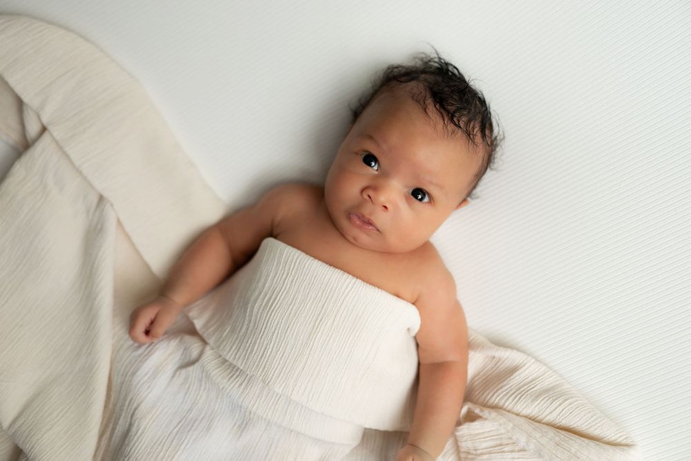 Newborn baby wrapped in a soft white swaddle blanket lying on a white background.