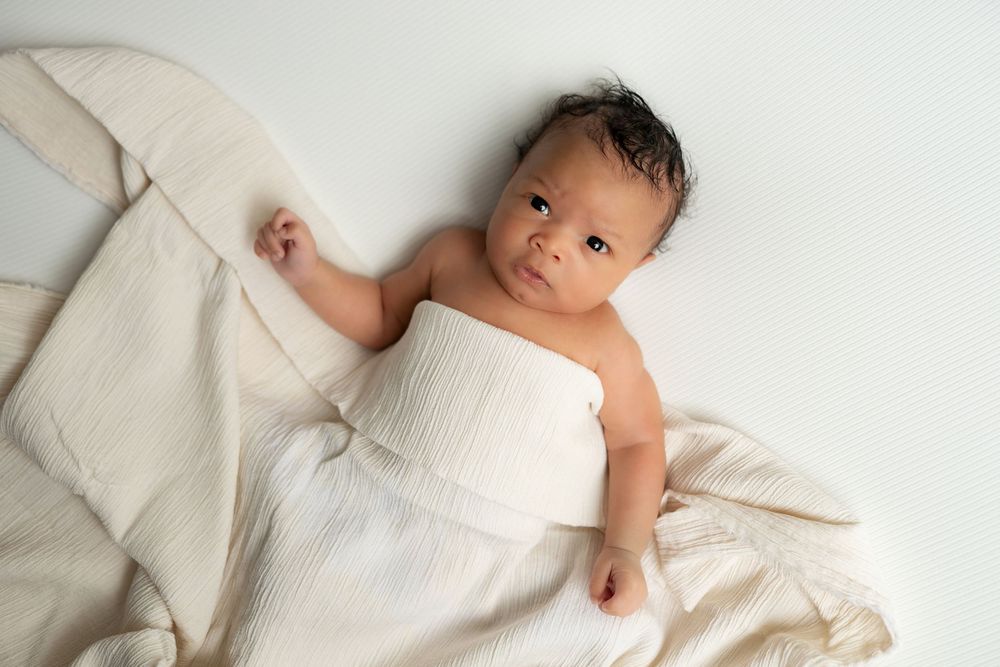 Newborn baby wrapped in white muslin cloth on cream textured blanket.
