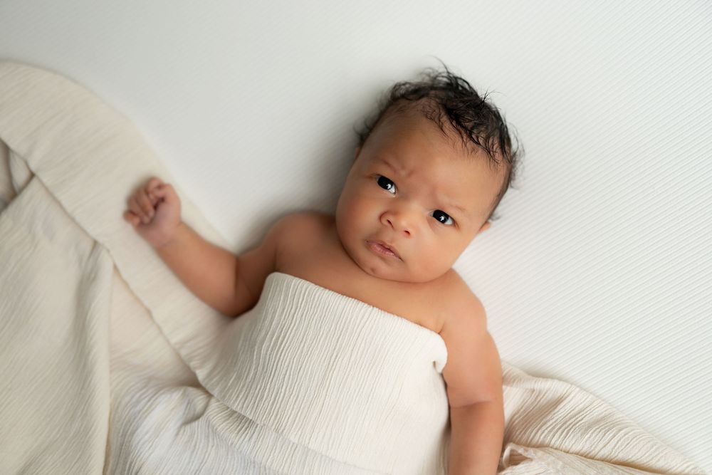 Newborn baby in white wrap lying on cream muslin blanket, looking up.