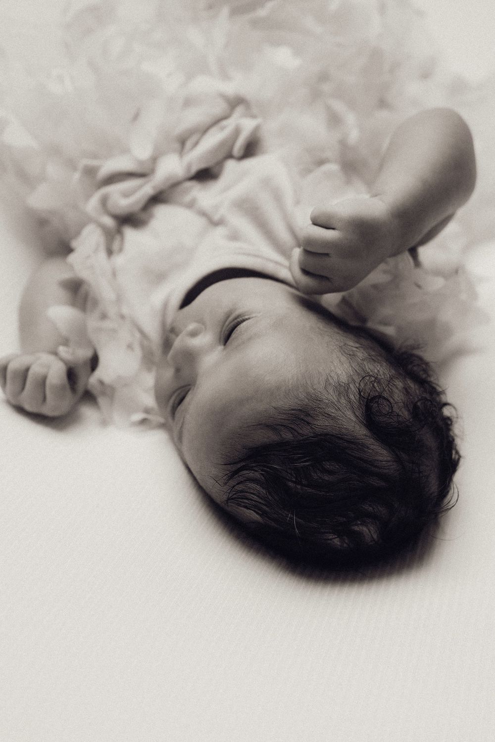 Black and white photo of sleeping newborn baby in ruffled white outfit lying down.