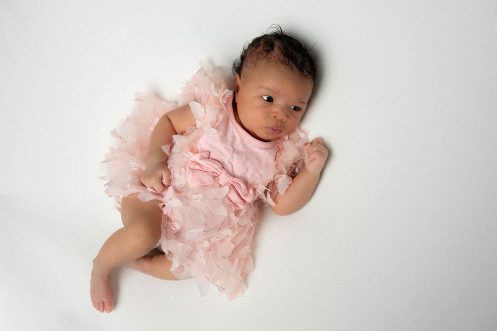 Newborn baby girl in a pink ruffled dress lying on a white background, looking up at the camera.