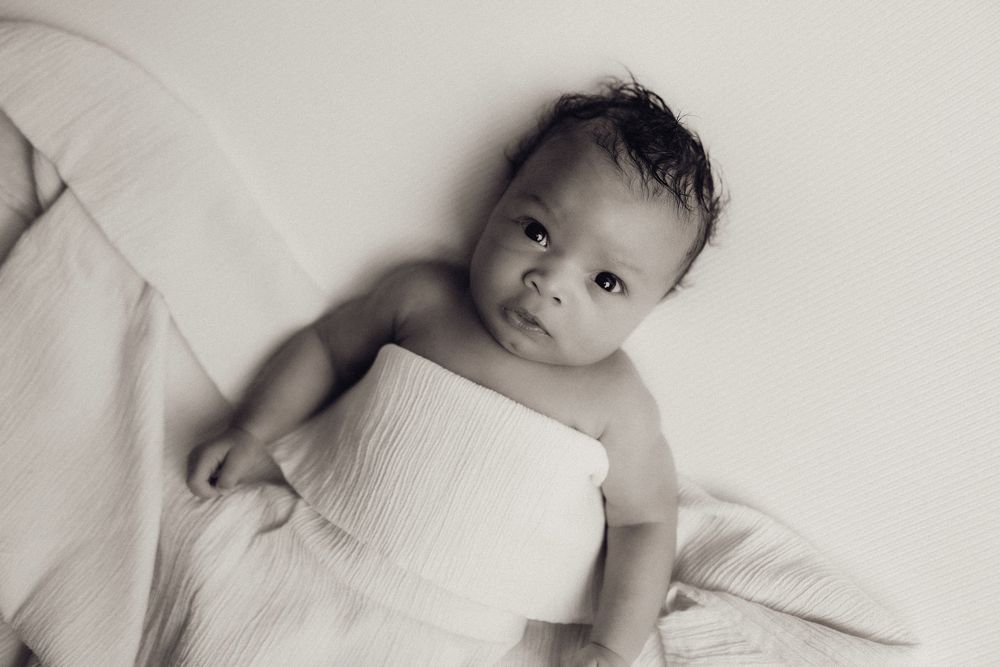 Black and white photo of newborn baby wrapped in white cloth lying on soft blanket.