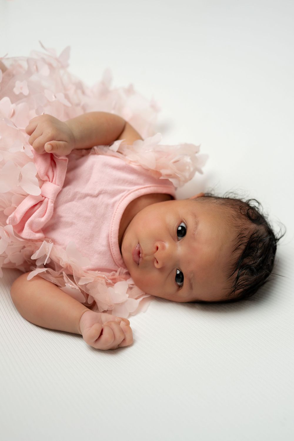 Newborn baby girl in pink ruffled dress lying on white background.
