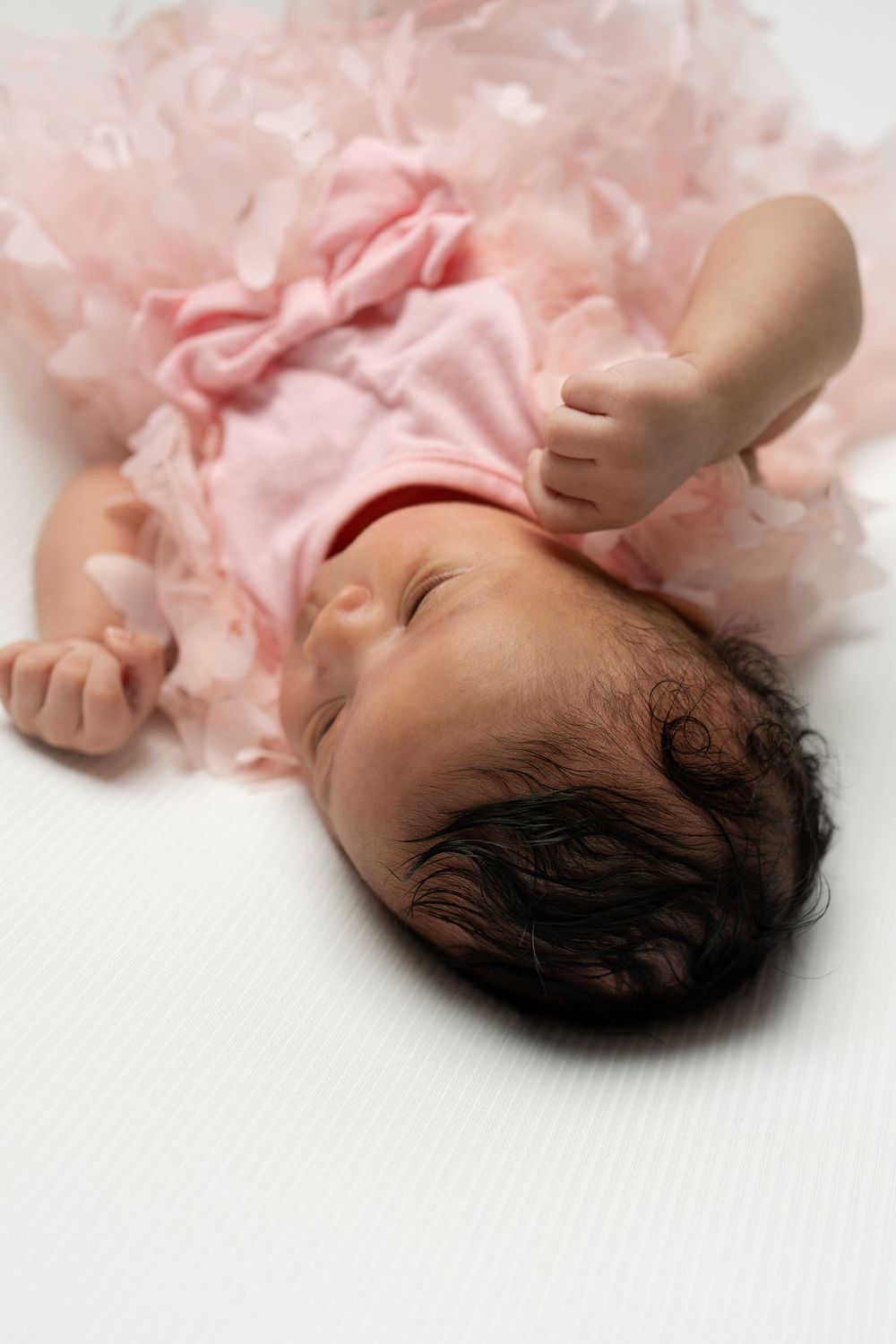 Newborn baby girl in a pink ruffled tutu lying on a white surface with dark hair and tiny fists raised.