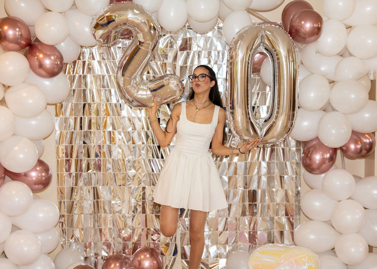 Young woman in white dress poses with gold number 20 balloons at a birthday party with balloon arch backdrop.