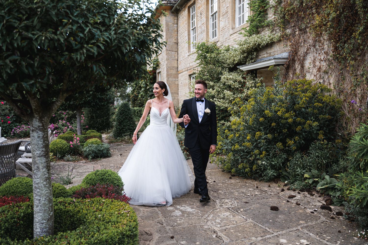 A bride and groom walk hand in hand along a stone path beside an ivy-covered manor house on their wedding day at Oxleaze Barn