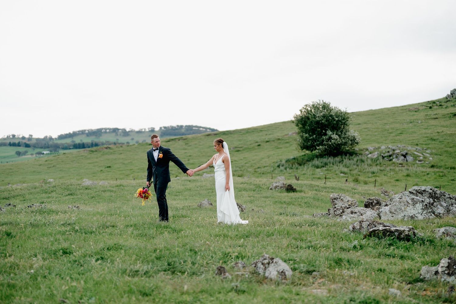 Bride and groom hold hands walking through lush green hillside meadow on their wedding day.