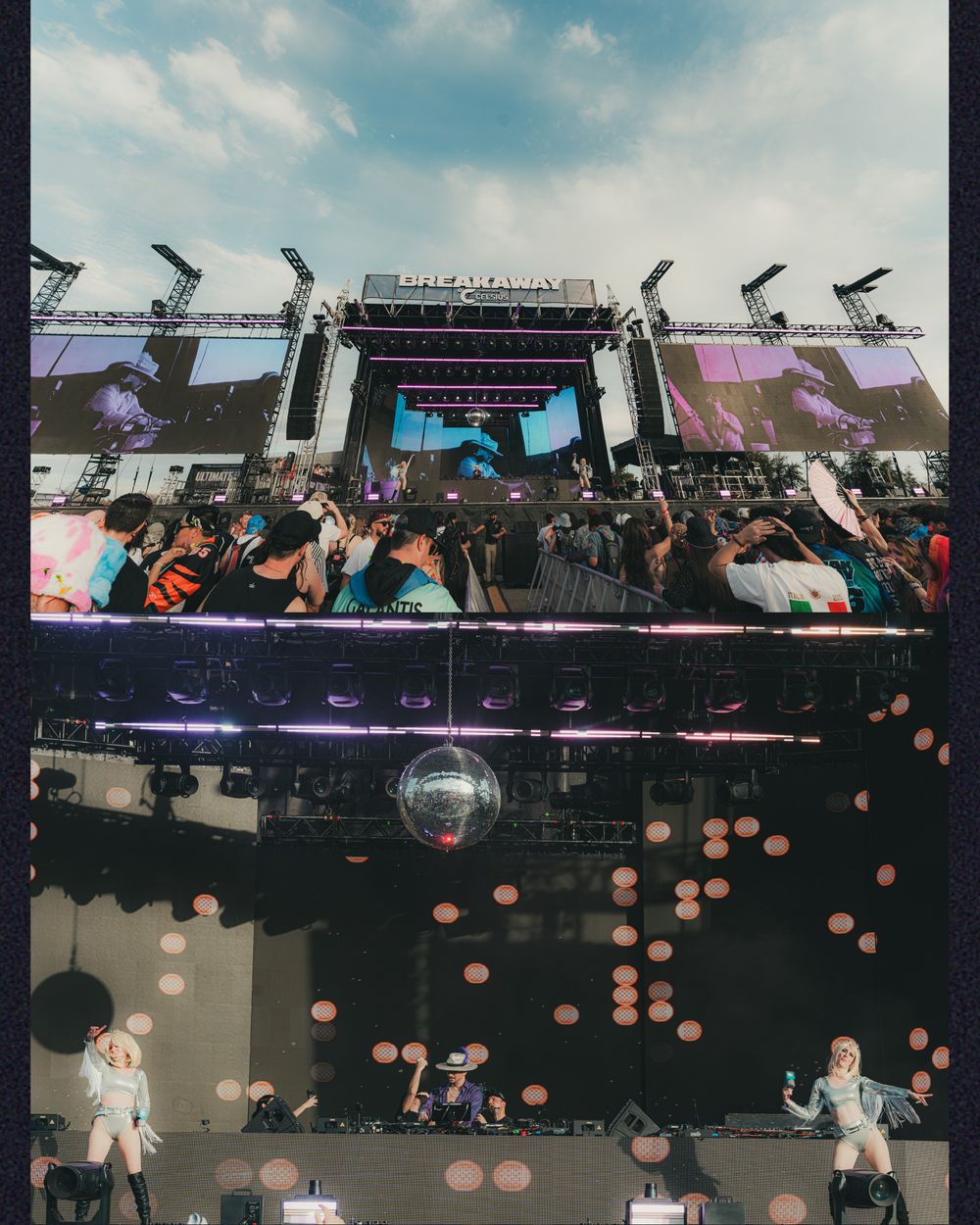 Breakaway music festival outdoor stage with large LED screens, crowd, and performers under a partly cloudy sky.
