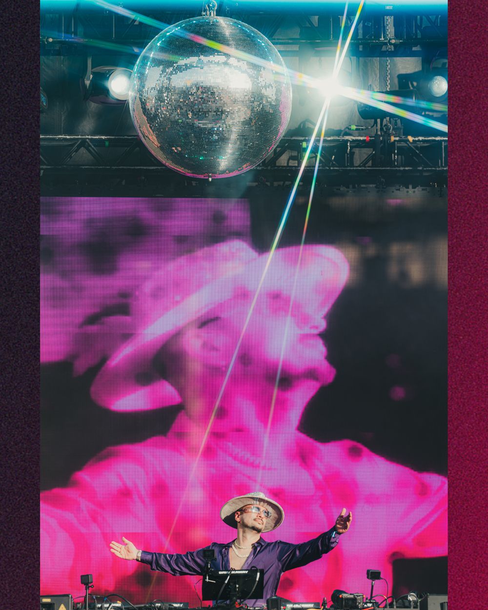 DJ performing under a glowing disco ball with vibrant pink LED screen backdrop at a live music event.