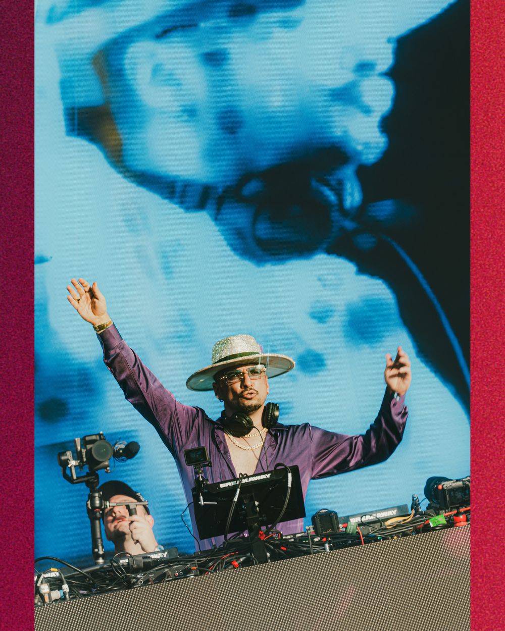 DJ in wide-brim hat raises hands behind turntables with blue projected backdrop at live music event.