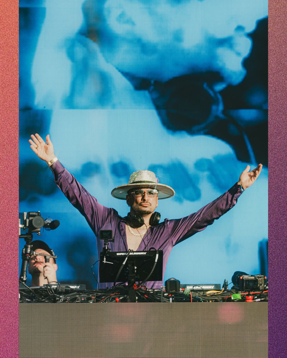 DJ in purple shirt and wide-brim hat raises arms triumphantly behind decks with blue projection backdrop.