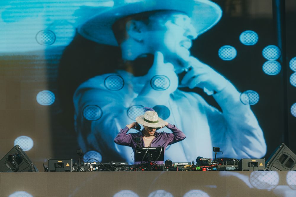 DJ in a wide-brim hat performing at a festival with a large projection screen behind them.