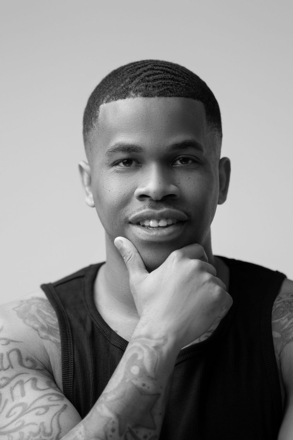 Black and white closeup portrait of a smiling person wearing a dark vest with visible arm tattoos.