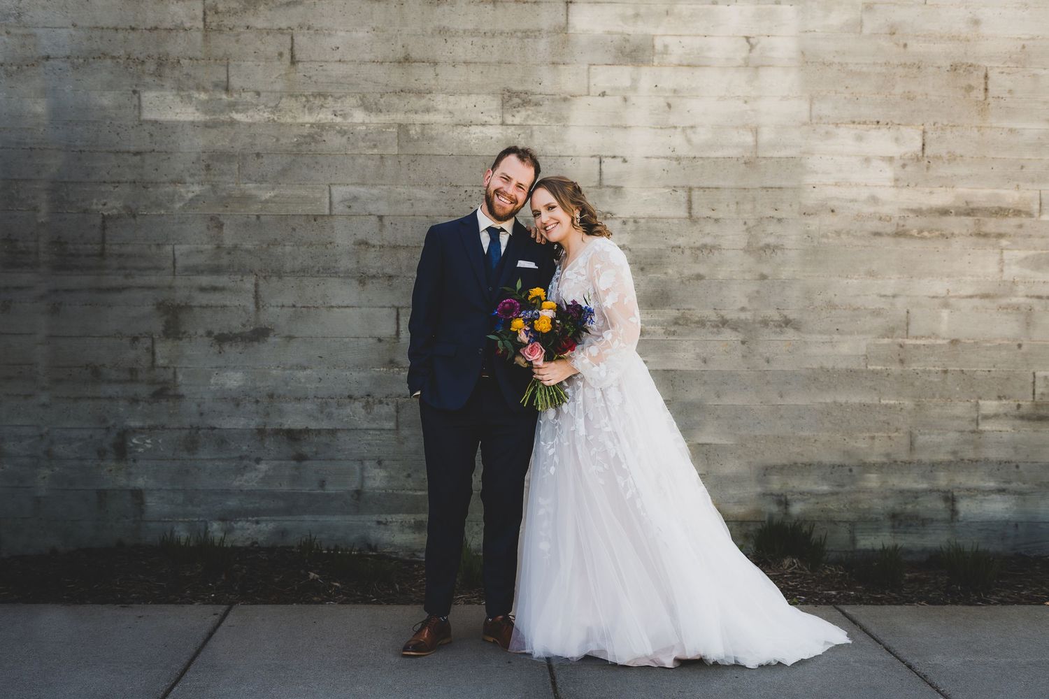 Newlyweds pose against stone wall with colorful bouquet and flowing white wedding dress.