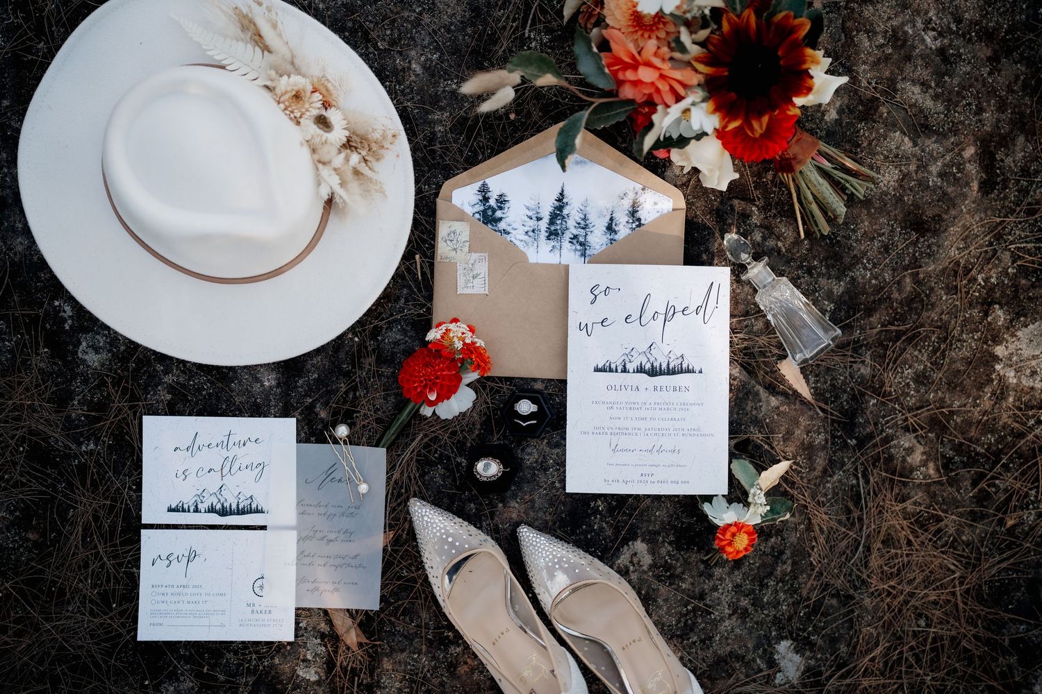 Elopement stationery flat lay featuring kraft envelopes, navy tree invitations, a white hat, heels, and red floral bouquet.