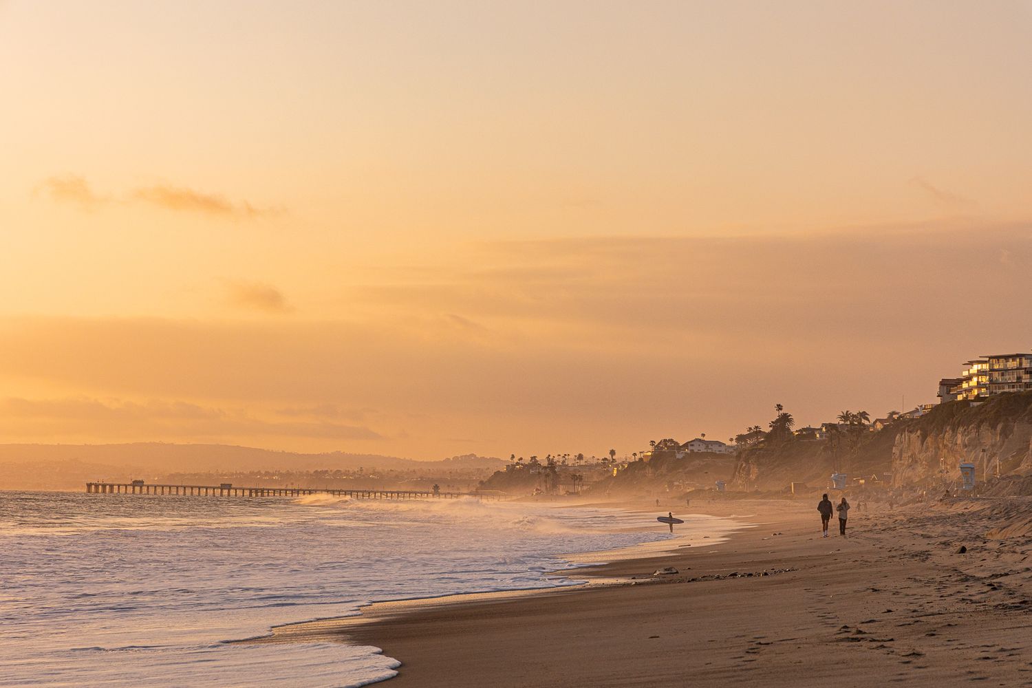 Golden sunset over a serene beach with couples walking along the shoreline near a pier in California.