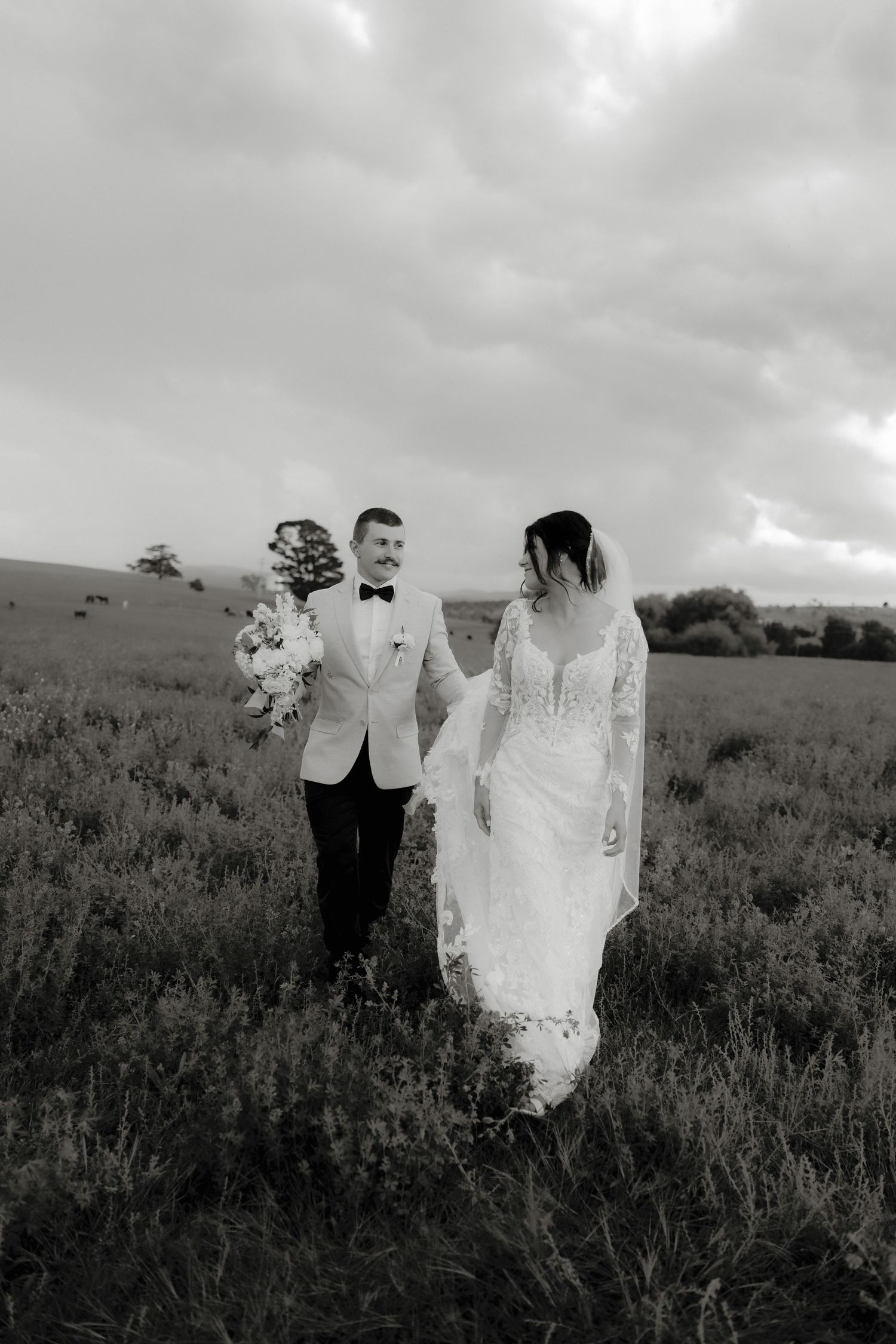 Black and white photo of a bride and groom walking hand in hand through a field on their wedding day.
