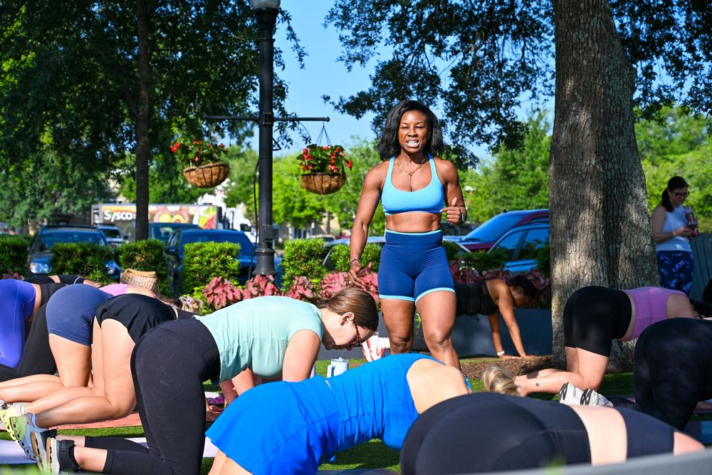 Fitness instructor leads an outdoor group workout class in a sunny park, wearing a blue sports bra and shorts.