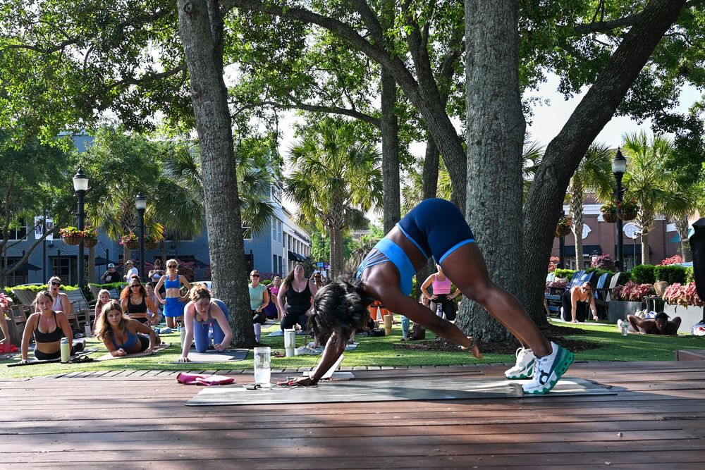 Outdoor yoga class in a park with participants in various poses on mats, surrounded by trees and spectators.