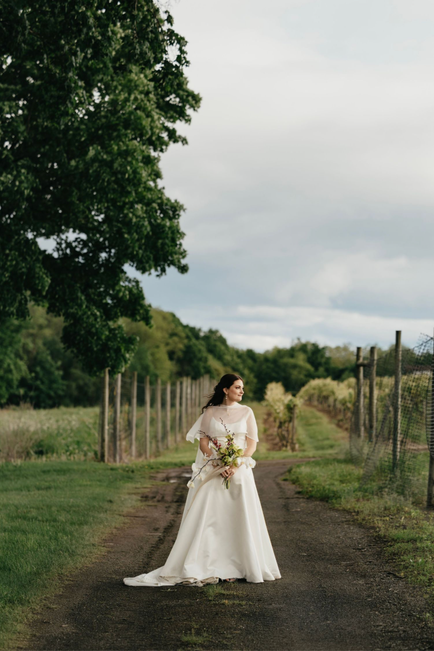Bride in white gown holding yellow bouquet stands on a rustic dirt path surrounded by lush greenery and vineyard fencing.