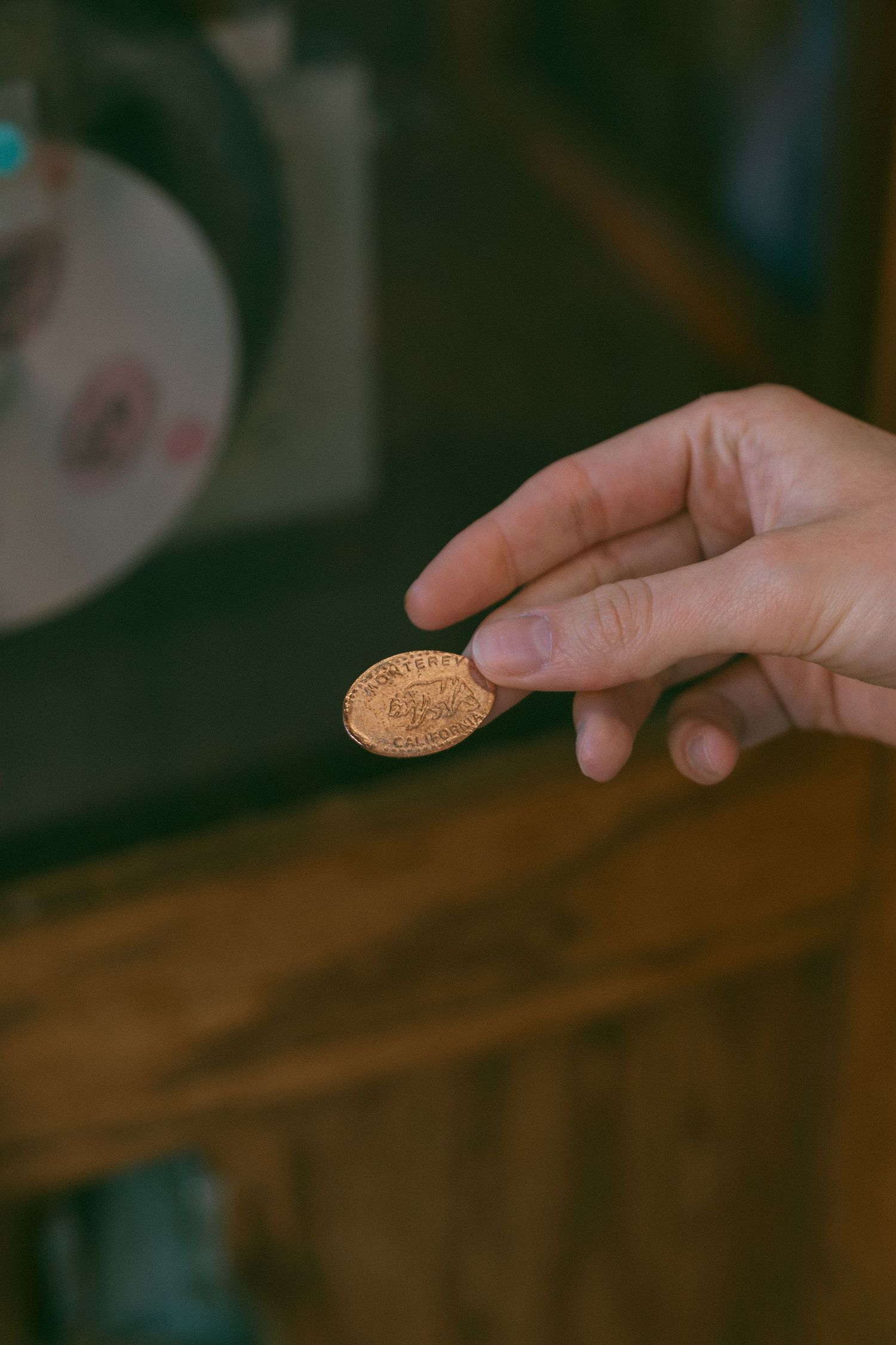 A hand holding a pressed penny souvenir coin, with a wooden background in a dimly lit setting.