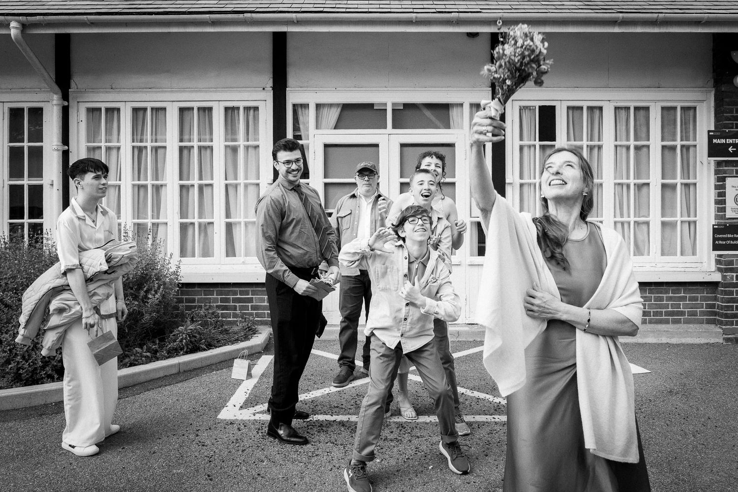 Black and white wedding photo of bride joyfully tossing bouquet outdoors with guests watching and reaching.