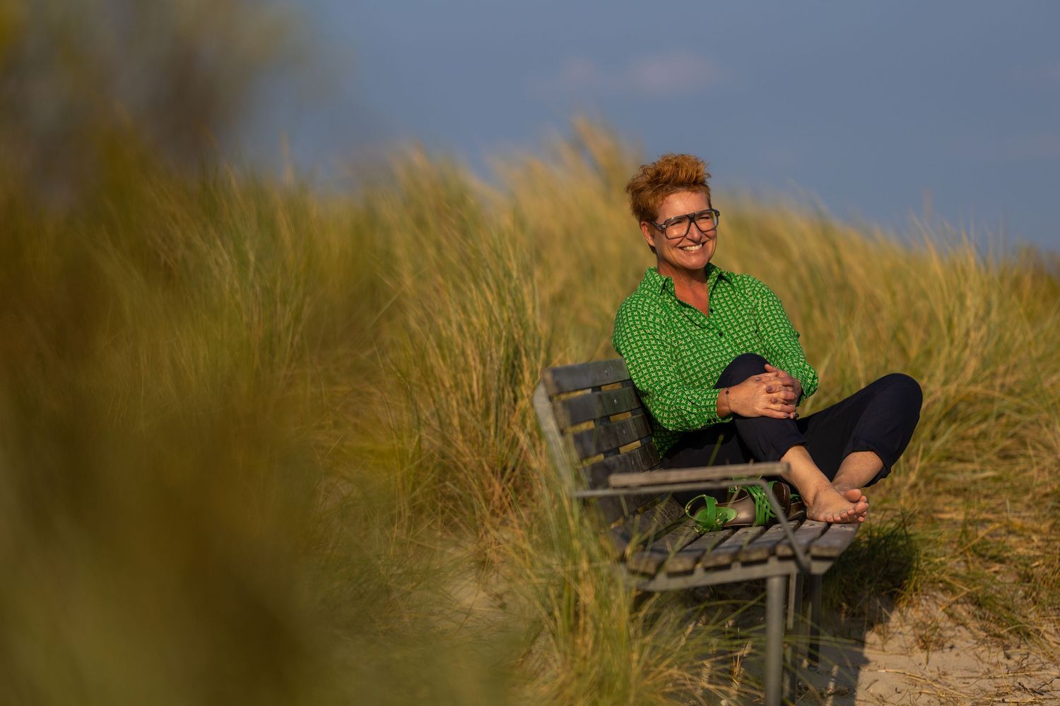 Person in green shirt sitting cross-legged on bench among sand dunes, smiling outdoors on sunny day.