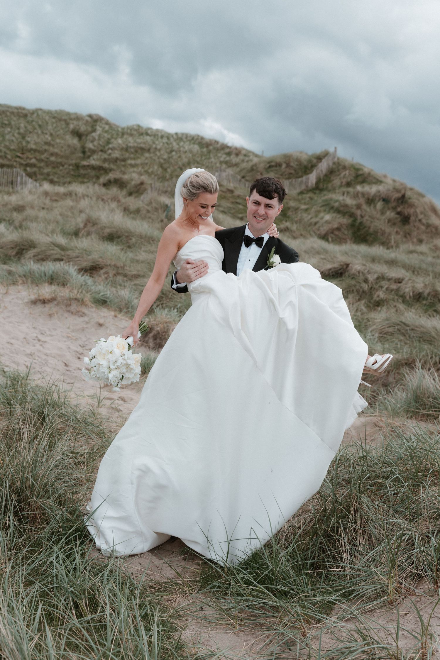 A bride and groom walk through sandy dunes, her white gown flowing in the wind as he holds her close on their wedding day.